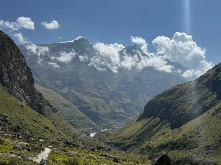 Trekkers on a path in the green valley above Mana with snow-capped peaks in the clouds ahead