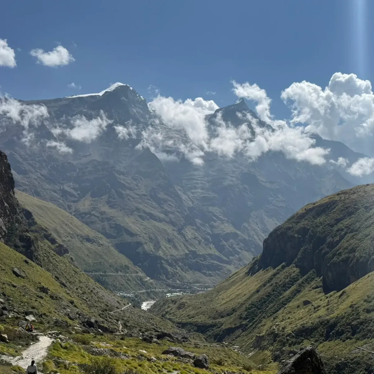 Trekkers on a path in the green valley above Mana with snow-capped peaks in the clouds ahead