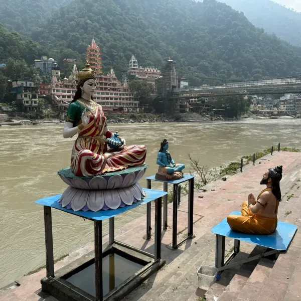 Statues of Mother Ganga, Shiva and a praying sage on the ghat at Rishikesh, with temples and a suspension bridge behind