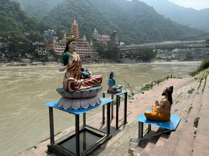Statues of Mother Ganga, Shiva and a praying sage on the ghat at Rishikesh, with temples and a suspension bridge behind