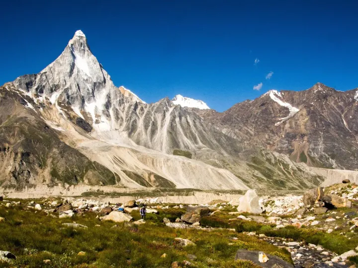 Mount Shivling seen from Nandanvan, a sharp pyramid of snow and rock above a green meadow with a lone trekker